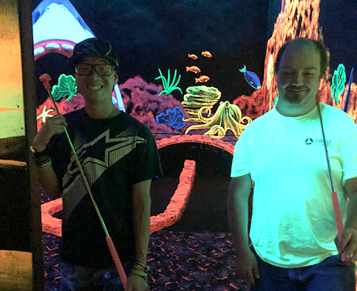 Two guys smile with their golf clubs in a game of black light mini golf.