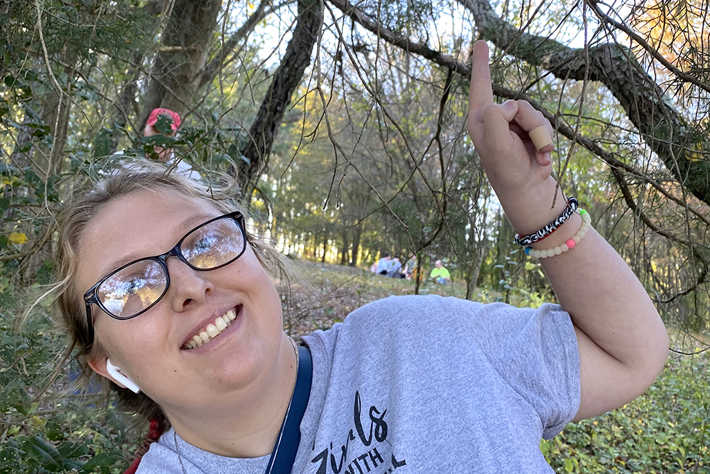 A girl smiles ouside in nature as she points up.